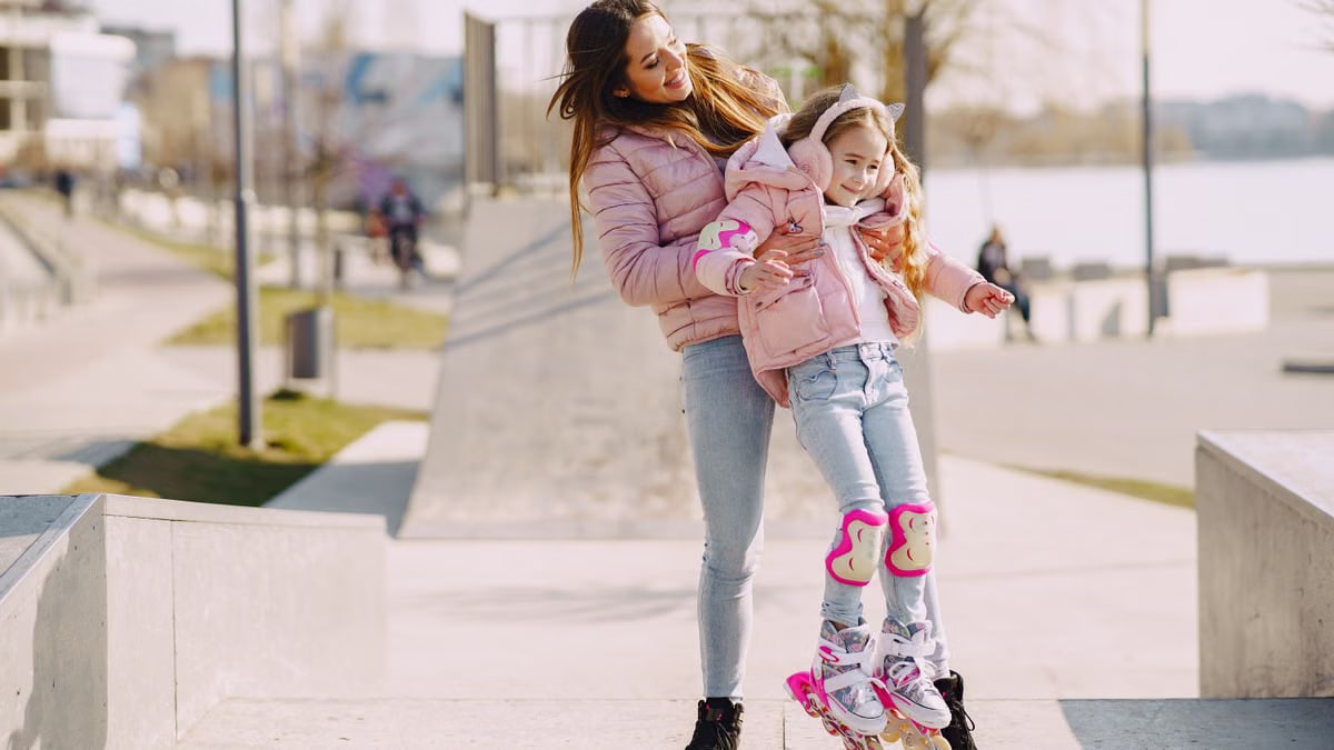Full body smiling mother carrying little girl wearing protection pads while roller skating on concrete ramp during active leisure in sunny spring day